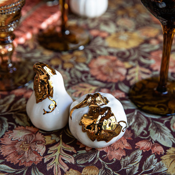 Two white ceramic pumpkin-shaped place card holders with gold accents from Juliska’s Forest Walk Tabletop Collection sit on a patterned floral tablecloth, next to dark-colored glassware.