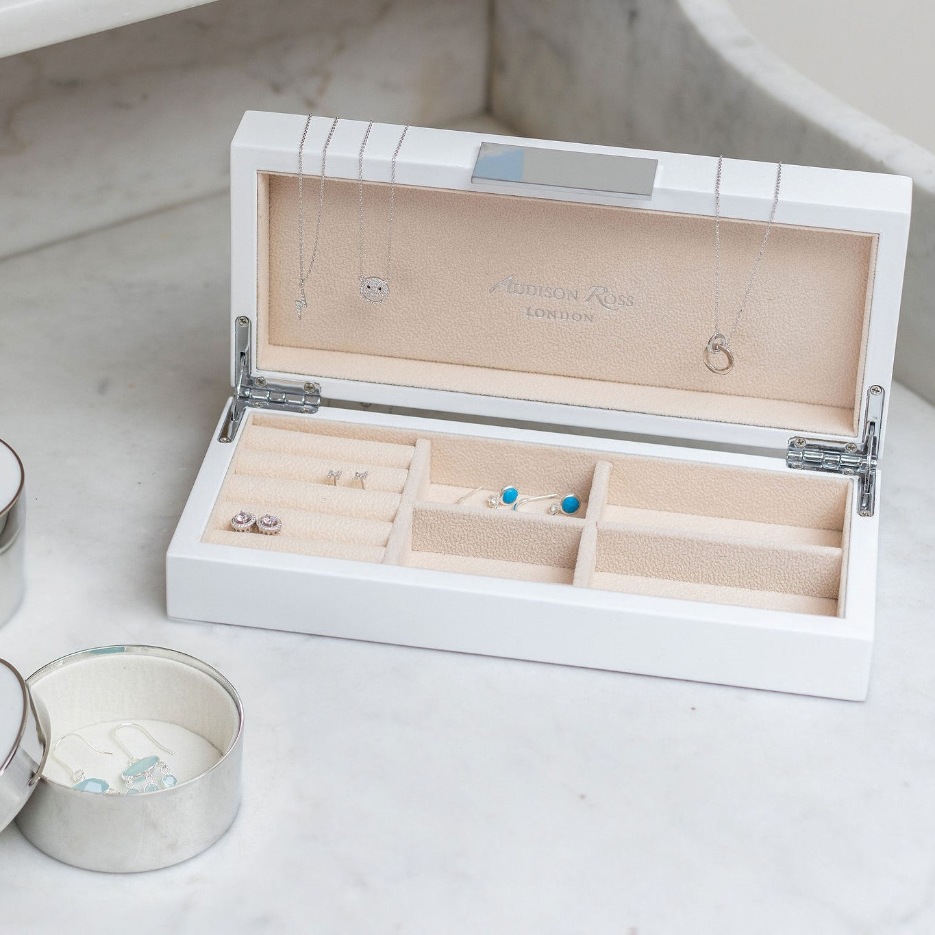 An Addison Ross White Lacquer Jewelry Box with a cream suede interior showcases necklaces, rings, and earrings elegantly displayed on a marble surface.
