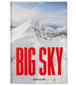 A book cover for "Big Sky" by Assouline shows a snowy Montana mountain under a blue sky, with bold red text reading "BIG SKY" and the brand name "Assouline" at the bottom.