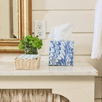 A Weezie Towels Marble Bath Collection tissue box with a blue and white swirl pattern sits on a marble counter beside a wicker basket and a small green potted plant in front of a mirror.