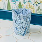 A hexagonal wastebasket from Weezie Towels’ Marble Bath Collection, made of recycled paper with a blue and white marbled pattern, rests on white tile near a wallpapered wall.