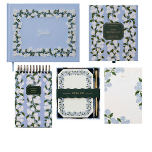 The Blue Hydrangea Desk Accessory Collection by Riffle Paper Co. features a hydrangea notepad, letter writing set with envelopes and pencil, journal, and memo pad—all adorned with elegant white and green floral designs.