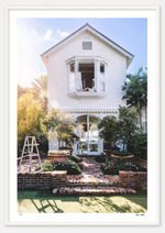 A two-story white house in West Palm Beach features an open second-story window. A person sits there, overlooking a brick pathway leading to the entrance marked by a "Please Sign In All Guests" sign. A ladder stands on the lawn beside the path, evoking the archival quality of Nick Mele Fine Art The Nest by Nick Mele Photography.
