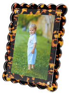 A young child in a light blue outfit stands on grass outdoors, pictured in Tara Wilson Designs' Scallop Frame Collection, featuring a scalloped, tortoiseshell-patterned border.