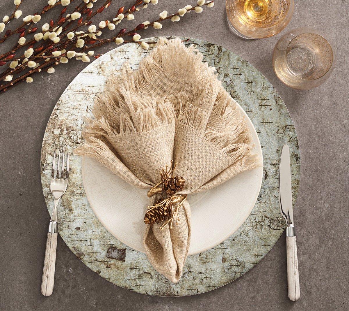 A table setting featuring the Kim Seybert Chalet Placemat, a white plate, fork and knife, beige fringed napkin tied with twine and pinecones, two glasses, and decorative pussy willow branches on a grey surface.