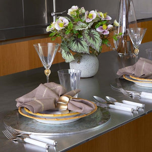 A modern table setting features Kim Seybert's Chalet Placemat, white and gold cutlery, beige napkins with gold rings, clear glassware, a floral arrangement, and nature-inspired decor on a metallic surface.