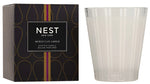 A Nest Moroccan Amber Candle from the Moroccan Amber Candle Collection is shown beside its black and gold box. The clear glass holder features subtle vertical stripes and contains a candle with patchouli notes.