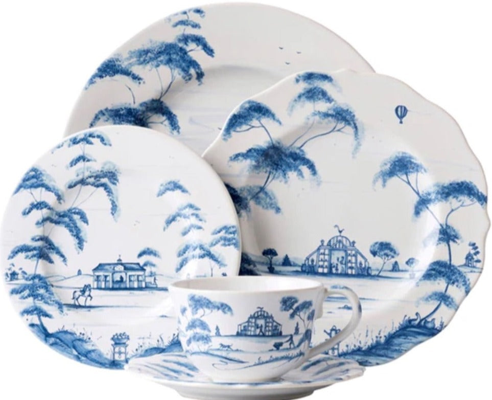 The Juliska Country Estate Delft Blue Dinnerware Collection features four white porcelain dishes and a teacup adorned with blue pastoral scenes of trees, houses, a horse stable, and a hot air balloon for countryside charm.