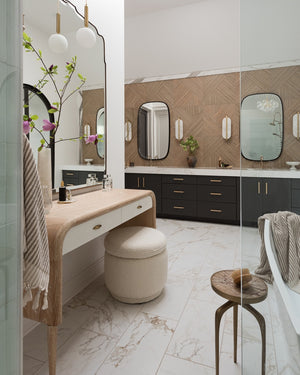 Modern bathroom featuring marble floors, a Made Goods Pierre Desk as a vanity, round stool, black cabinets, large rounded mirrors, decorative wall panels, and contemporary lighting fixtures.