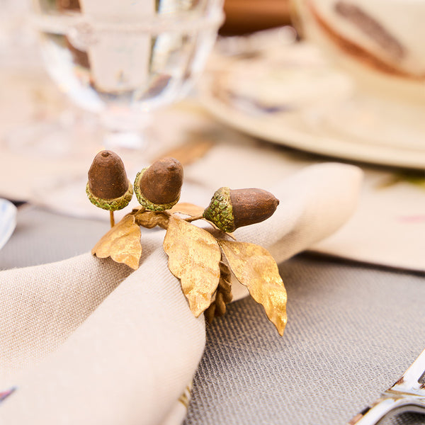 A beige napkin from Juliska's Forest Walk Tabletop Collection is held by a gold ring with leaf and acorn accents, adding elegant oak-inspired decor to the dining table.
