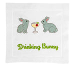 The August Morgan Drinking Bunny Cocktail Napkins feature two embroidered gray rabbits facing a cocktail glass with a cherry, framed by classic hemstitching. "Drinking Bunny" is stitched in green for a playful touch.