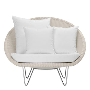 The Lounge Chair in Schooner Snow by Janus et Cie, featuring a round wicker design with a white cushion and three white throw pillows on metal legs, adds midcentury modern style to any luxury space.