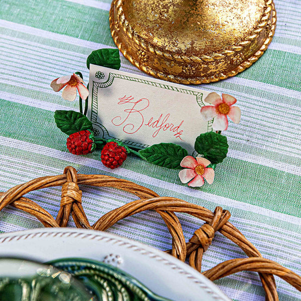 The Meadow Walk Raspberries Place Card Holder Set by Juliska features a holder decorated with artificial berries and flowers, ideal for charming table settings on striped linens beside woven placemats and gold accents.