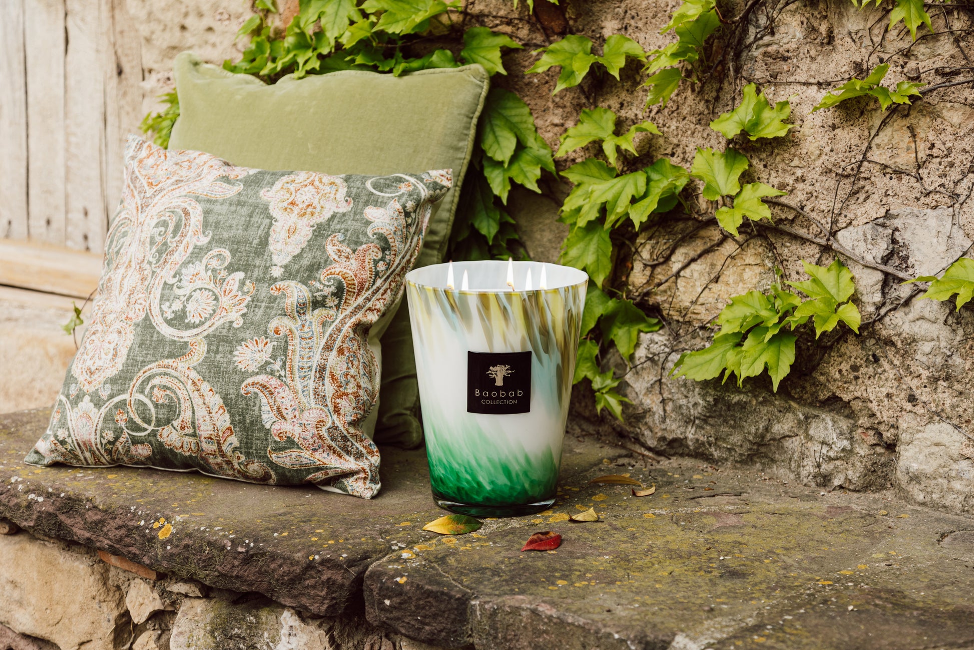 A decorative Baobab Collection Eden Forest candle in a green and white holder, next to a paisley pillow on a stone bench, surrounded by ivy.