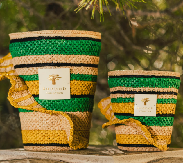 Two Baobab Collection Fety Milalao Candles, in decorative green, black, and yellow striped woven baskets with a gold "Baobab" label and ylang-ylang scent, are placed on a stone surface outdoors against blurred greenery.