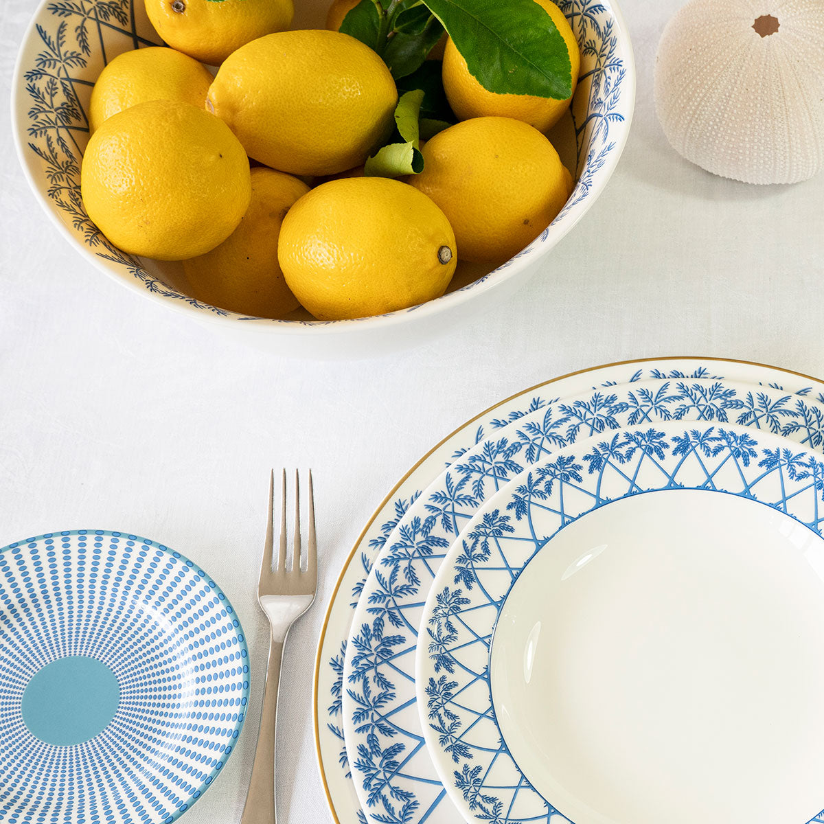 A white table set with the Azure Blue Dinnerware Collection by Pink House Mustique, complete with a bowl of lemons, small dish, fork, and white decor—capturing elegance with a hint of Exotic Tropics flair.