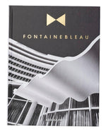 The Assouline book "Fontainebleau" features a black-and-white cover image of architectural beauty, adorned with gold lettering. It exudes luxury, reflecting the grandeur reminiscent of the iconic Fontainebleau hotel.