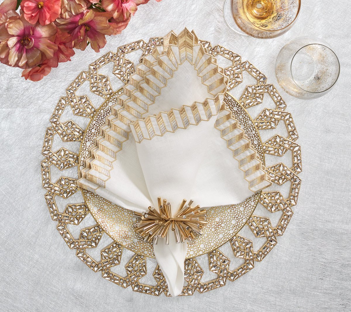 A white napkin with gold trim, folded decoratively and held by the Kim Seybert Fringe Napkin Ring, rests on a gold geometric charger beside a glass and pink flowers for a glamorous look.