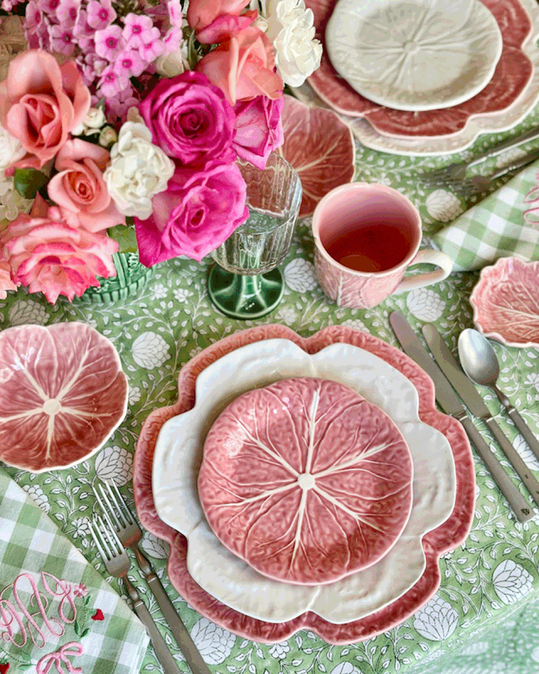 A floral-themed table is set with Bordallo Pinheiro’s Pink Cabbage Leaf Dinnerware and Serveware Collection, silver cutlery, a pink mug, glass, and a vase of pink flowers atop a green patterned tablecloth.