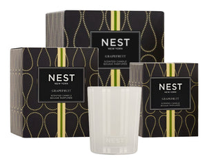 A clear glass candle from the Nest Grapefruit Candle Collection, exuding a fresh scent, is showcased in front of three elegant black and gold Nest boxes.