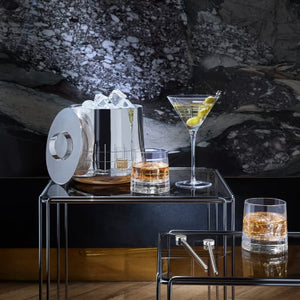 A Christofle Graphik Insulated Ice Bucket sits with tongs, a martini glass with olives, and two double-insulated whiskey glasses on nested glass tables against a dark, textured background.