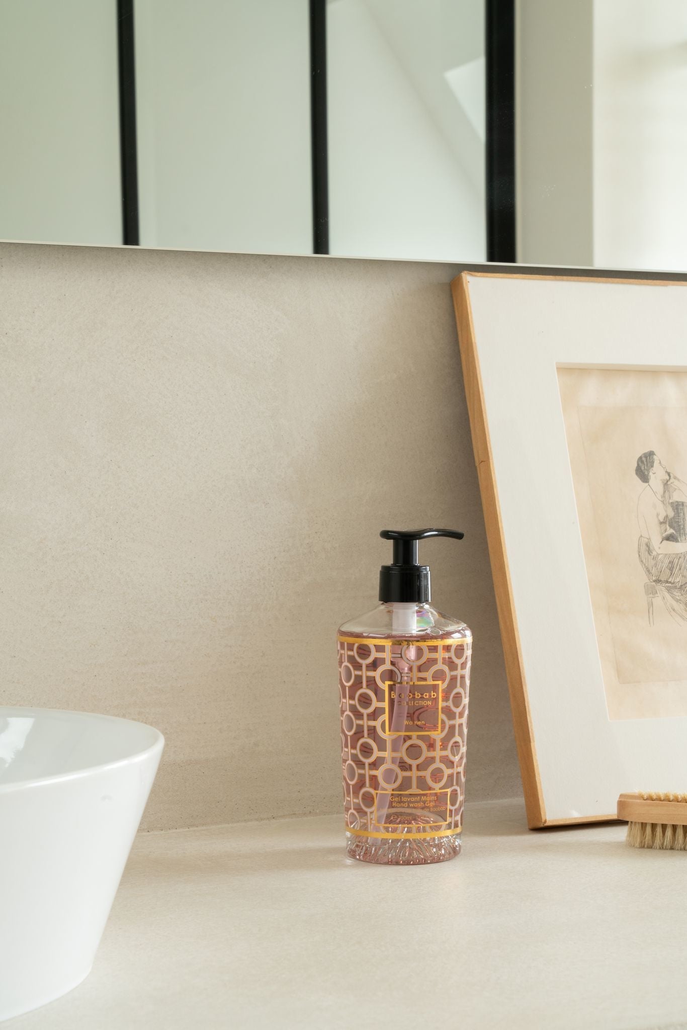 The Baobab Collection Women Body Wellness Collection soap dispenser by Baobab adds a floral touch to the bathroom countertop, set beside a framed sketch and white sink, with a mirror in the background.