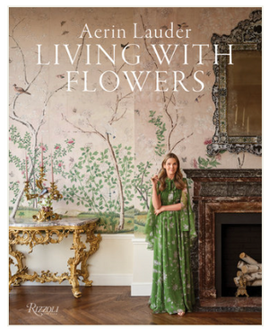 A woman in a green dress stands by a fireplace in an elegant room with floral wallpaper and ornate furniture, featuring the Rizzoli book "Aerin Lauder: Living With Flowers," an inspiring guide to entertaining with beautiful blooms.