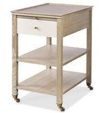 The Alexander Narrow Bedside Table by Highland House features a blonde cerused finish, raised edge top, single white drawer with brass handle, two open shelves, and four caster wheels for stylish bedside storage.