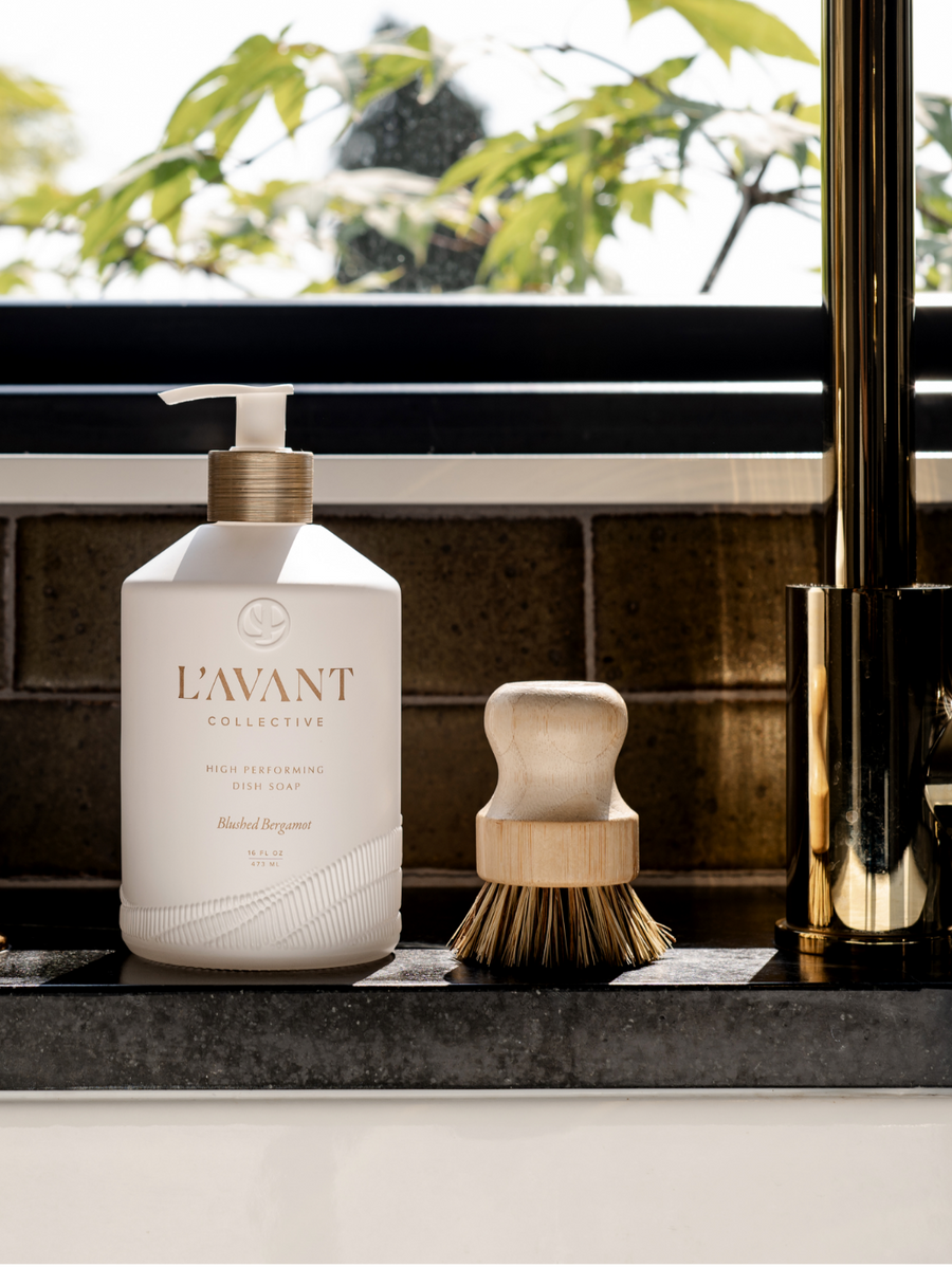 A bottle of L’Avant Collective Blushed Bergamot Dish Soap and a wooden scrub brush rest on a kitchen counter by the sink and gold faucet, sunlight streaming in as hints of their signature scent fill the air.