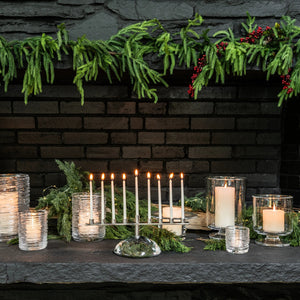The Simon Pearce Riverstone Menorah with lit candles rests on a stone mantel adorned with greenery, red berries, and white candles in glass holders.