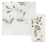 The Holly & Sprig Linen Collection by Kim Seybert features white linen napkins and placemats with embroidered green leaves, pine needles, red berries, and gold accents—ideal for a festive Christmas table setting.