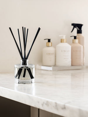 A Blushed Bergamot Reed Diffuser by L'Avant Collective sits on a marble countertop, with soap and lotion dispensers and a spray bottle in the background.