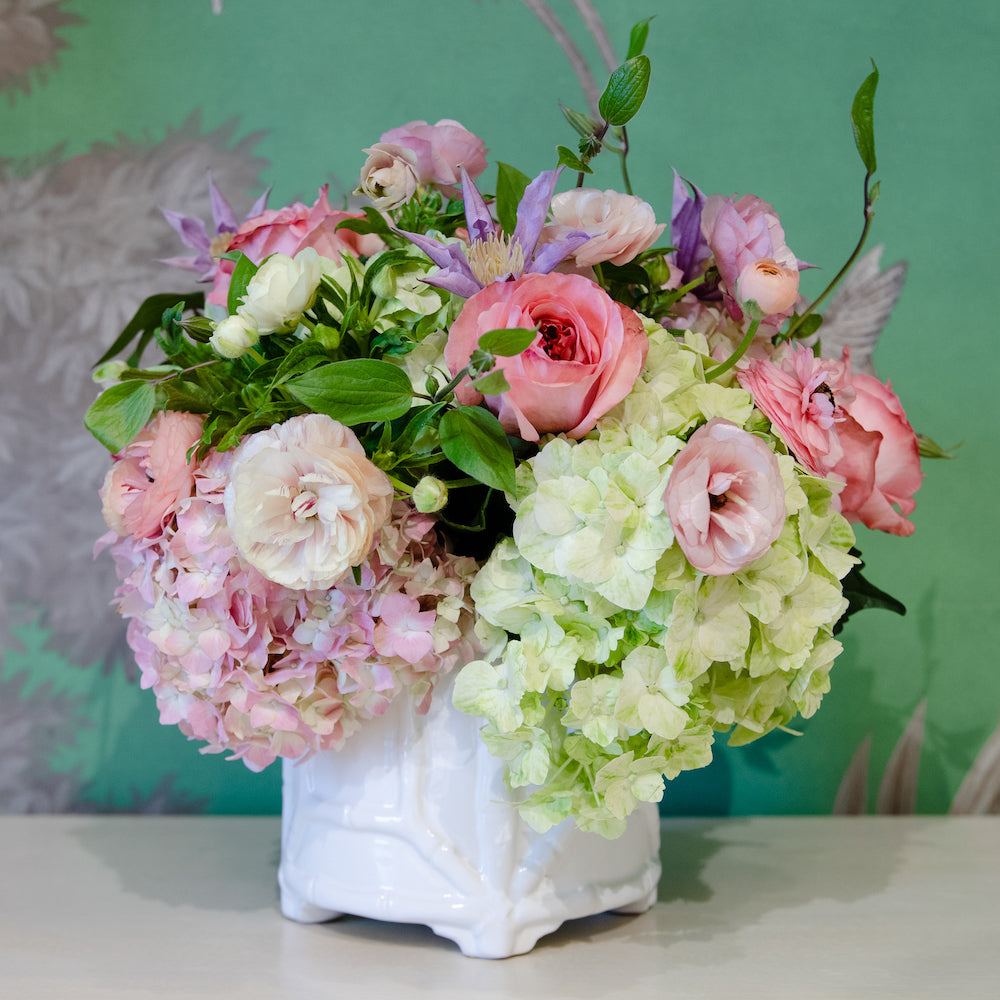 The Live Designer's Choice Floral Arrangement by Hive Floral Studio features a hand-picked bouquet of mixed flowers and greenery in a white vase, beautifully set against a green background.