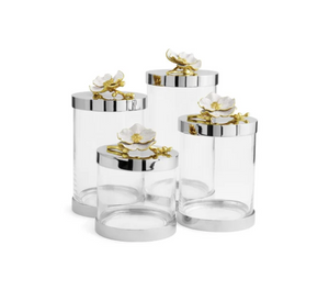 The Michael Aram Orchid Canister set features four clear glass canisters with silver lids, each adorned with a white and gold flower accent, adding the pure elegance of the Orchid Collection to any space.