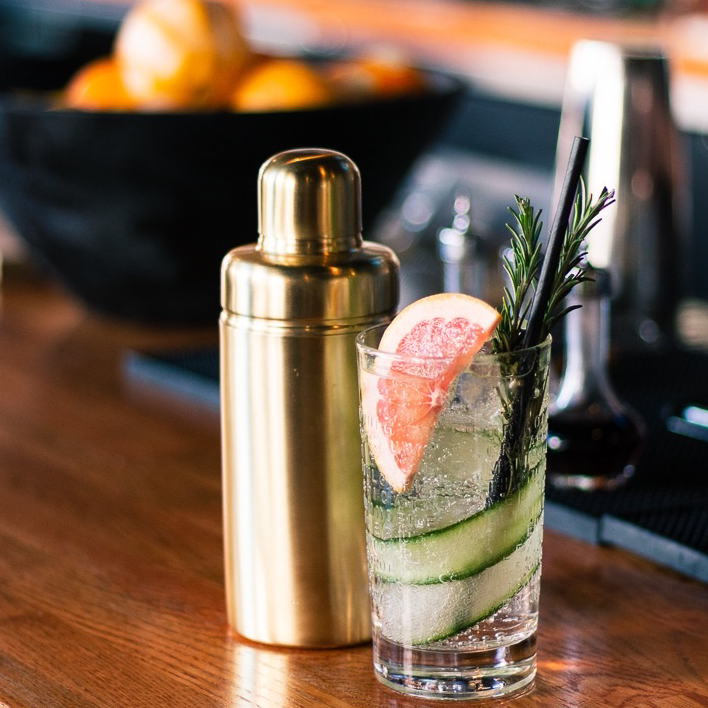 A clear cocktail with cucumber slices, grapefruit, and rosemary sits beside the Be Home Luxe Verona Cocktail Shaker on a wood bar—ideal for an ethically crafted cocktail event.