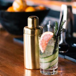 A clear cocktail with cucumber slices, grapefruit, and rosemary sits beside the Be Home Luxe Verona Cocktail Shaker on a wood bar—ideal for an ethically crafted cocktail event.