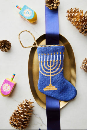 The Bauble Stockings Light the Menorah Stocking by Bauble Stockings features a blue menorah design on a gold platter, elegantly adorned with a blue ribbon. This heirloom-quality stocking is beautifully surrounded by pinecones and colorful dreidels on a white background, setting an ideal scene for honoring family traditions.