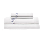 A neatly stacked set of John Robshaw’s Stitched Indigo Bedding Collection—white organic cotton sheets with blue stitched detailing—displayed against a plain white background.