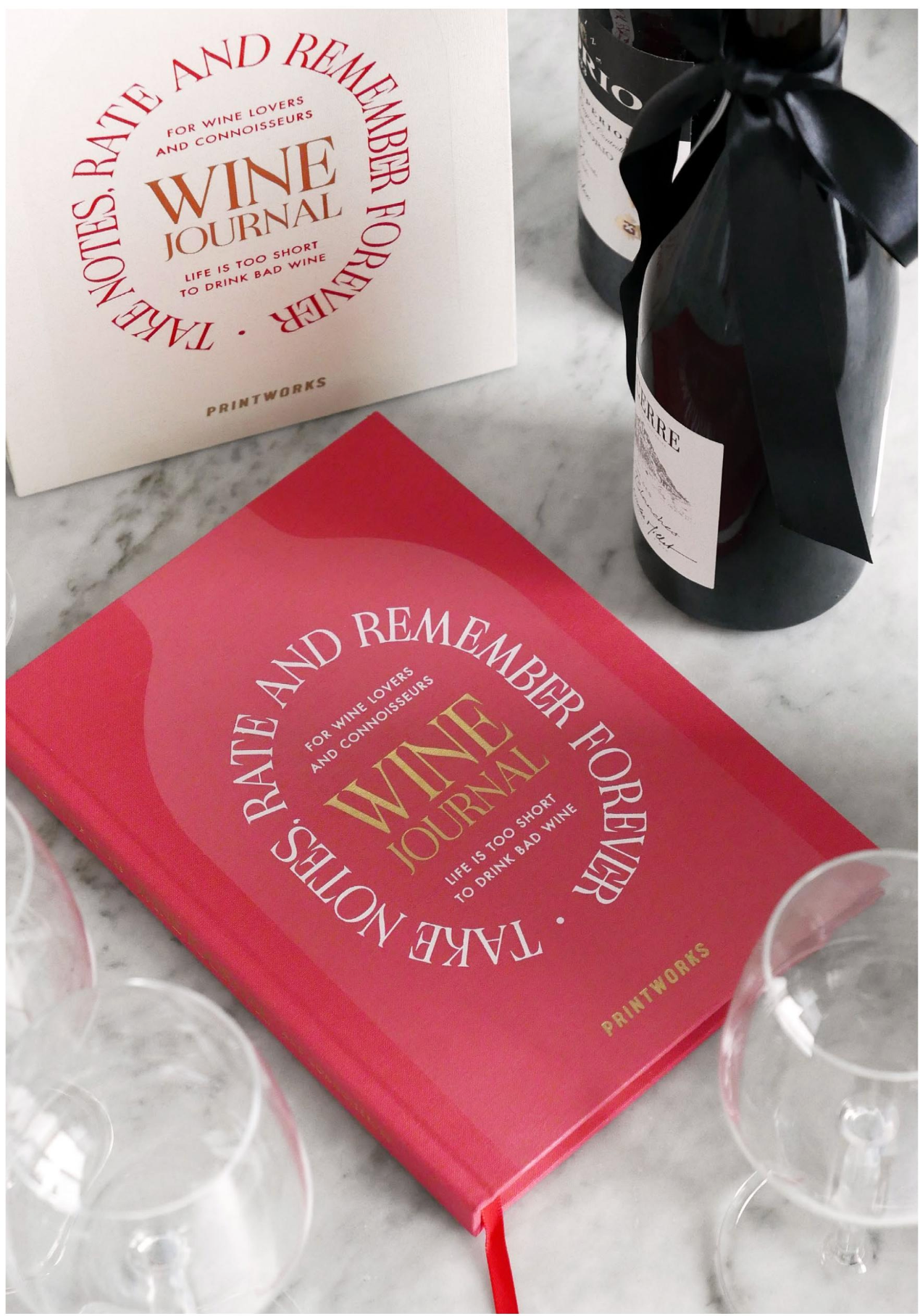 The PrintWorks Wine Tasting Journal rests on a marble surface with wine glasses, a ribboned wine bottle, and its packaging—ideal for logging tasting notes or updating your sweetness chart.