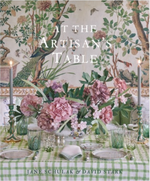 Book cover for "At The Artisan's Table" by Jane Schulak & David Stark, from Vendome, features a floral centerpiece, green checkered tablecloth, candles, and a decorative wall with birds and trees—celebrating contemporary tabletop artisans.