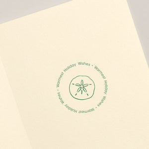 The Crane Seashells Wreath Card Set features a cream holiday card with a green sand dollar illustration at the center, encircled by the phrase "Warmest Holiday Wishes" for an elegant coastal touch.