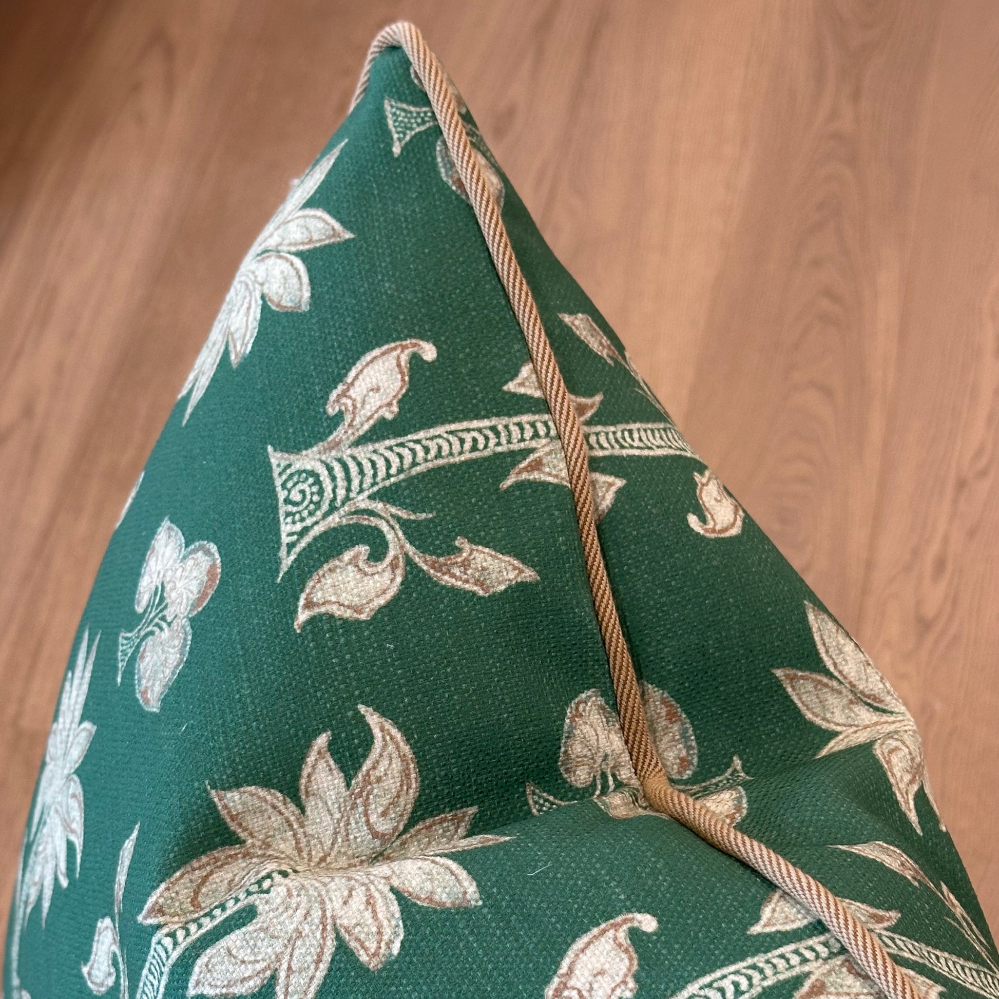 The Associated Design Mazari Palm Outdoor Pillow, featuring a green fabric with beige floral pattern and beige piping, is displayed on a wooden floor.