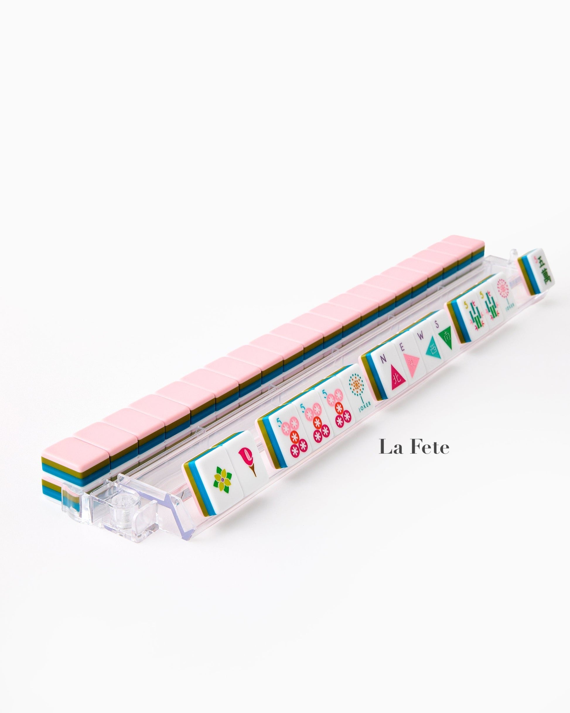 A row of hand-painted Mahjong tiles from the Oh My Mahjong Tile Collection is arranged on a clear rack, displaying vibrant designs. "La Fete" is shown beside the rack on a white background.