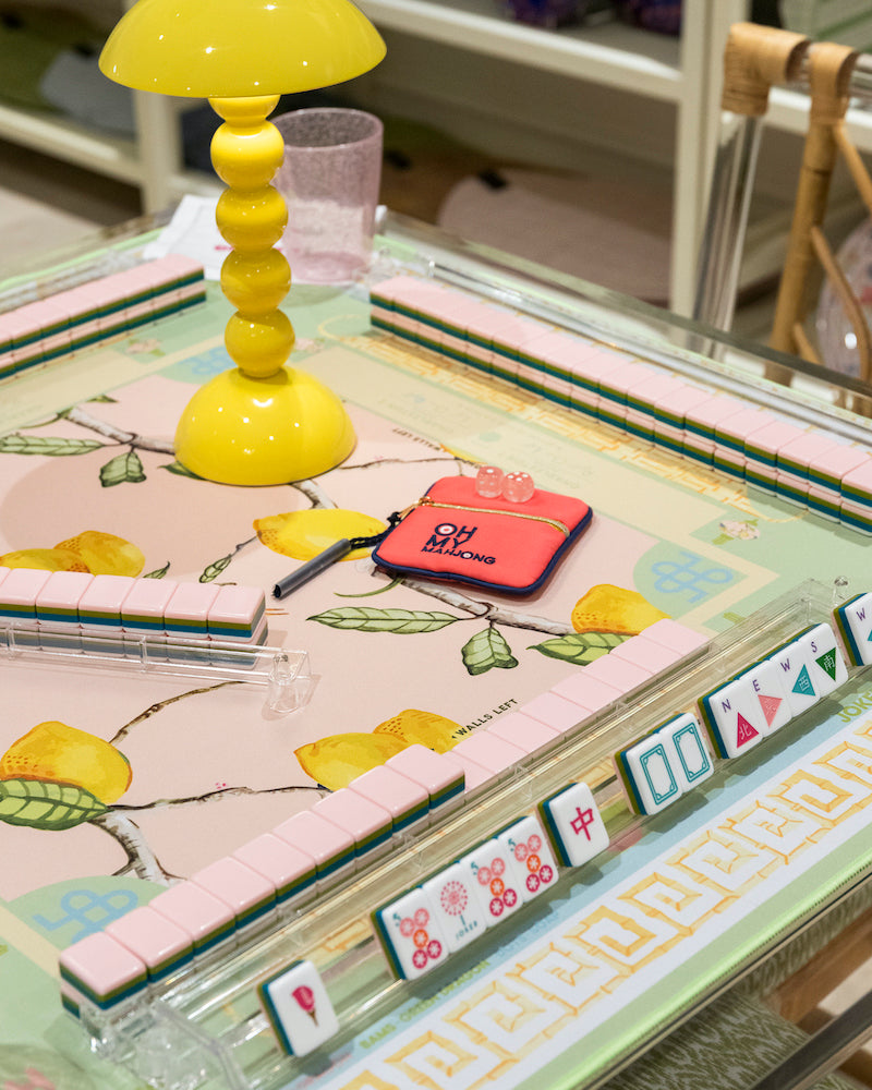 The Oh My Mahjong Tile Collection delivers a vibrant Mahjong gaming scene on a lemon-patterned table, featuring hand-painted tiles, racks, a yellow lamp, pen, red pouch, and glass.