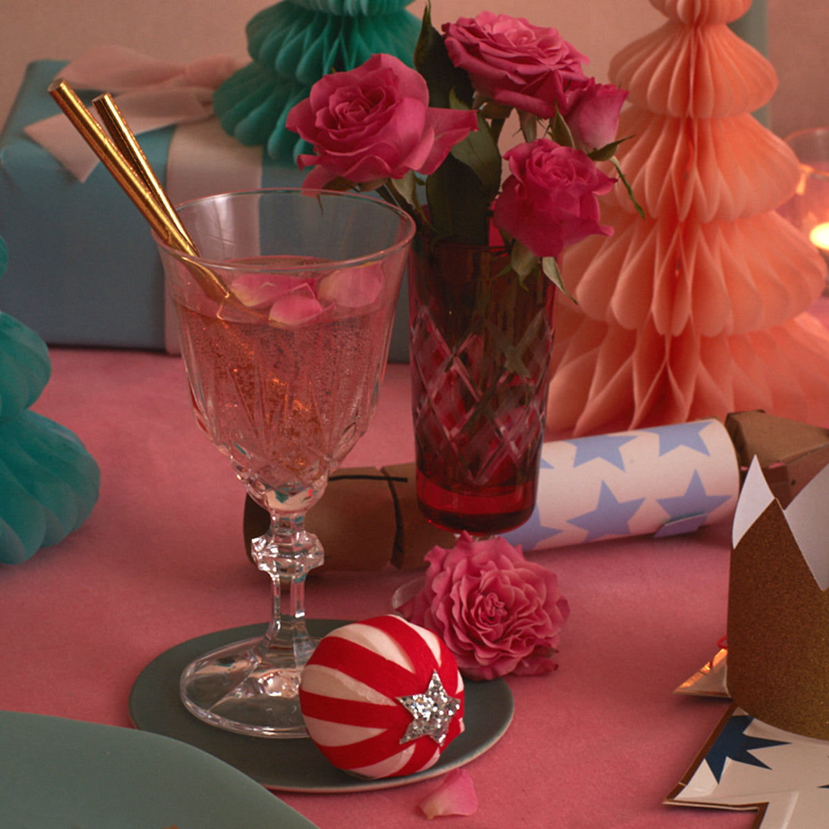 A decorated table with Peppermint Candy Surprise Balls by Meri Meri, a vase of pink roses, wrapped gifts, party crackers, a crown, and festive decor on a pink cloth—perfect for holiday surprises or stocking fillers.