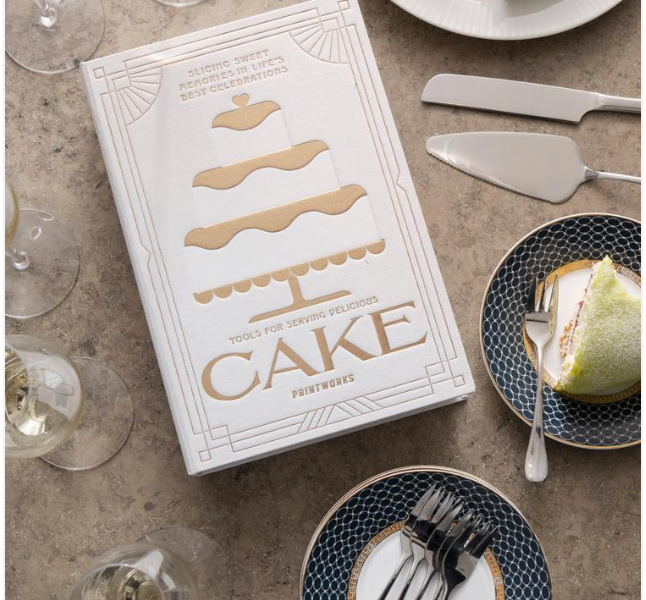 A white PrintWorks "The Essentials Cake Tools" boxed set sits on a table with cake servers, plates of cake, forks, and champagne glasses—both stylish kitchen decor and the perfect tools for cake serving.