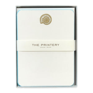The Nautilus Note Card Box Set by Printery includes ecru cards with azure blue edges and a gold nautilus shell design, wrapped with a paper band branded "Printery Oyster Bay New York.