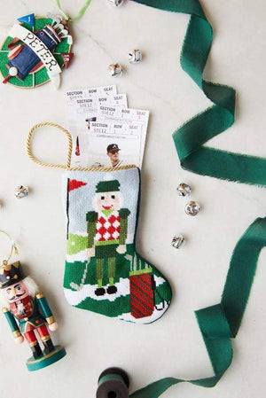 The Bauble Stockings Christmas Birdie Nutcracker Needle Point Stocking holds show tickets and is surrounded by ornaments, a nutcracker, green ribbon, and silver jingle bells on a white surface.