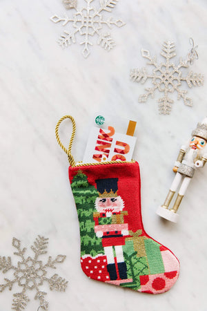 The Bauble Stockings Classic Nutcracker Needle Point Stocking holds a gift card, surrounded by three silver Christmas ornaments and a small nutcracker figurine on a white surface.