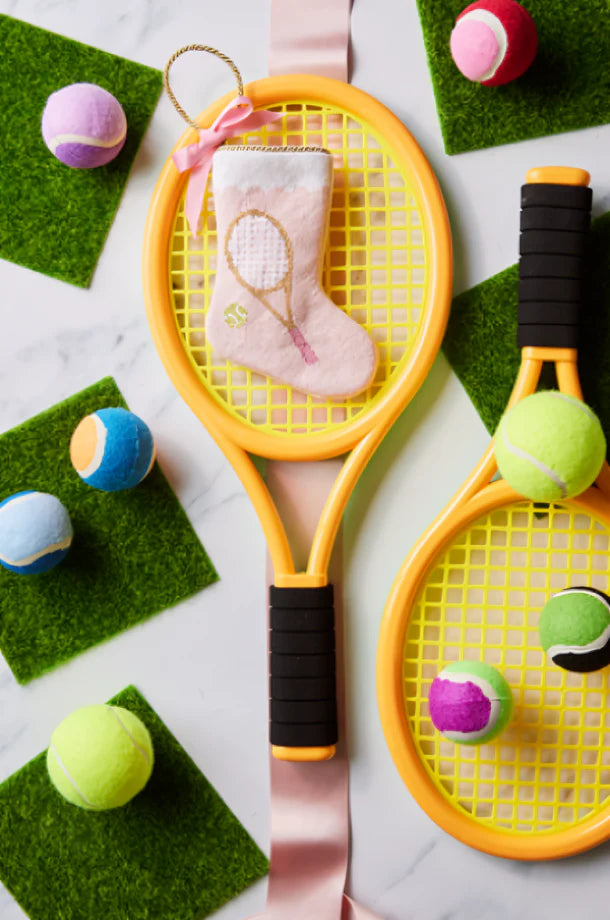 Two yellow toy tennis rackets, colorful toy balls, and green felt squares rest on a white surface. Adding a festive touch is the Bauble Stockings Tennis Pink Needle Point Stocking, perfect for playful holiday decor.